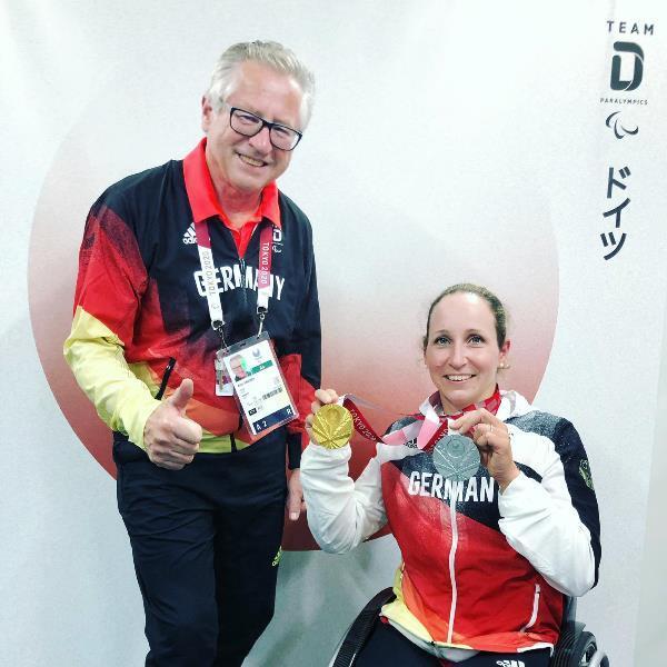 Zwei Personen in Sportkleidung zeigen stolz eine Gold- und eine Silbermedaille vor einem Hintergrund mit Team-Logo.