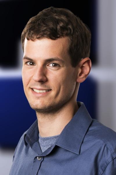 Portr&auml;t eines jungen Mannes mit kurzem Haar, der in einem blauen Hemd l&auml;chelt, vor einem verschwommenen Hintergrund.