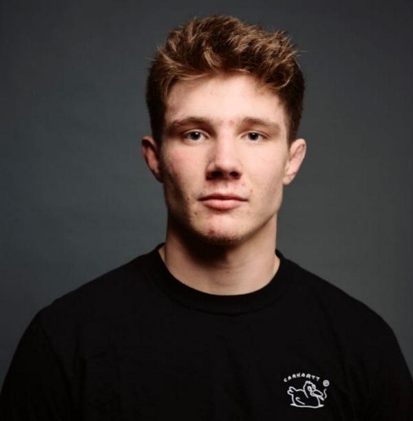 Portr&auml;t eines jungen Mannes mit kurzer Frisur, der einen schwarzen T-Shirt tr&auml;gt, vor einem grau-blauen Hintergrund.