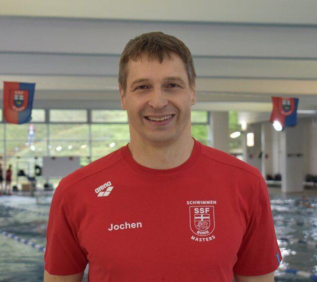 L&auml;chelnder Mann in rotem Schwimmsportshirt steht in einem Schwimmbad mit blauen Wasserbahnen im Hintergrund.