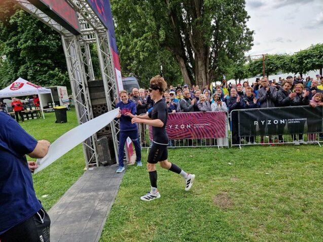 Sportler &uuml;berquert die Ziellinie, w&auml;hrend Zuschauer applaudieren. Im Hintergrund sind Werbebanner sichtbar.