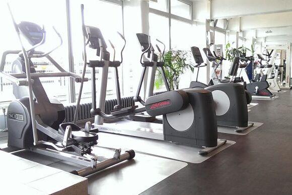 Modernes Fitnessstudio mit mehreren Crosstrainern und Laufb&auml;ndern entlang gro&szlig;er Fenster. Pflanzen im Hintergrund.