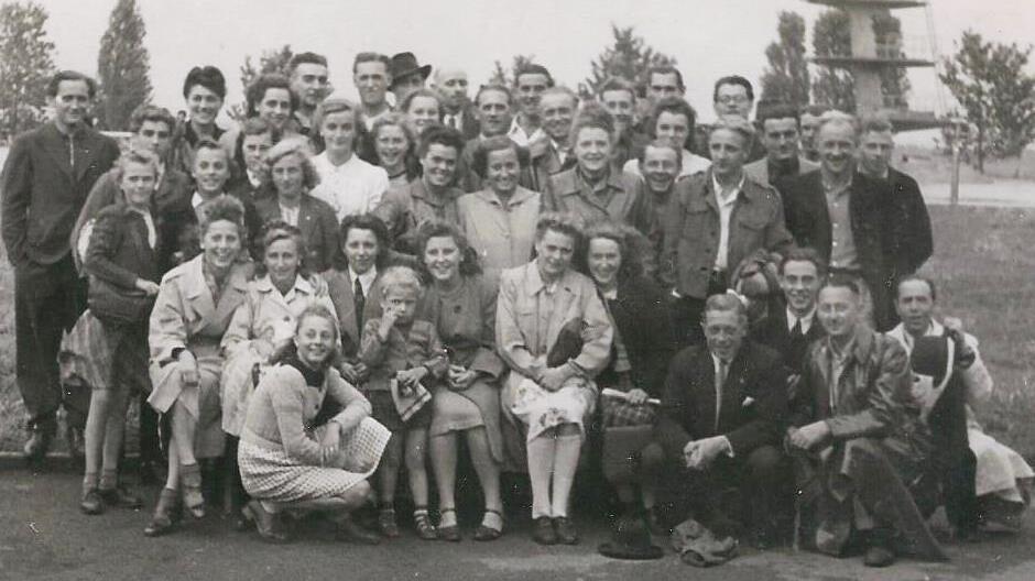 Gruppenselfie von etwa 50 Personen im Freien, umgeben von B&auml;umen und einem modernen Geb&auml;ude im Hintergrund.