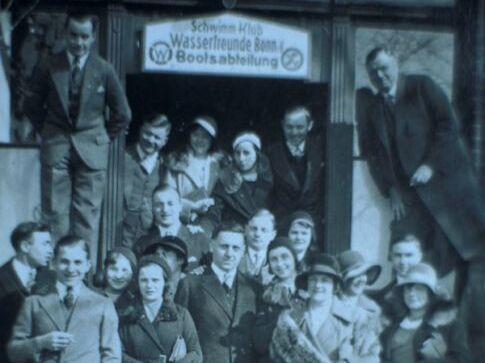 Gruppenfoto vor einem Vereinsgeb&auml;ude mit mehreren M&auml;nnern und Frauen in eleganter Kleidung und H&uuml;ten.
