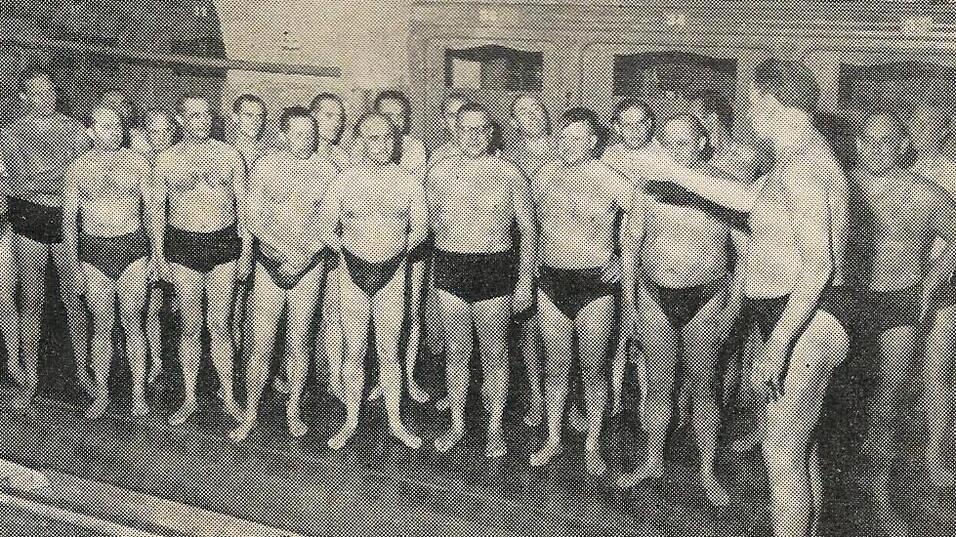Gruppe von M&auml;nnern in Badehosen steht in einem Schwimmbad, ein Mann spricht zur Gruppe.