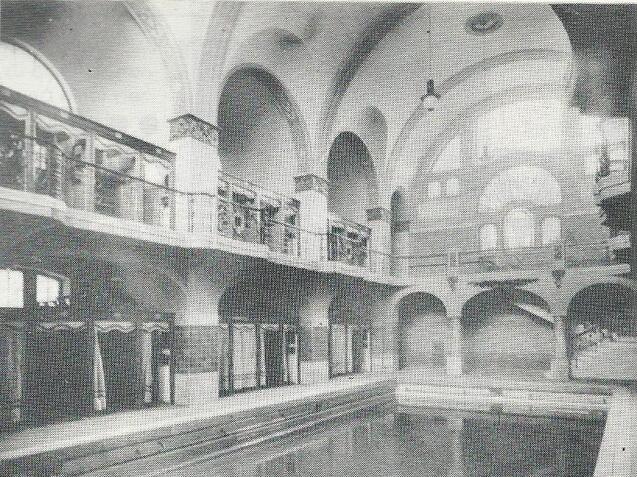 Historische Schwimmhalle mit gro&szlig;en B&ouml;gen, Balustraden und einem leeren Schwimmbecken im Erdgeschoss.