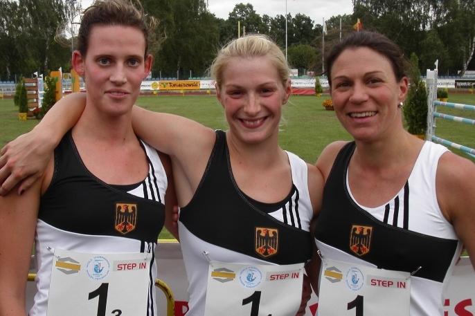 Drei Athletinnen in Sportkleidung stehen eng beieinander und l&auml;cheln vor einer Wettkampfst&auml;tte f&uuml;r Modernen F&uuml;nfkampf.