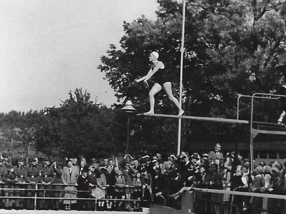 Eine Springerinnen steht auf einem Sprungbrett &uuml;ber einem Schwimmbecken, w&auml;hrend Zuschauer am Rand beobachten.