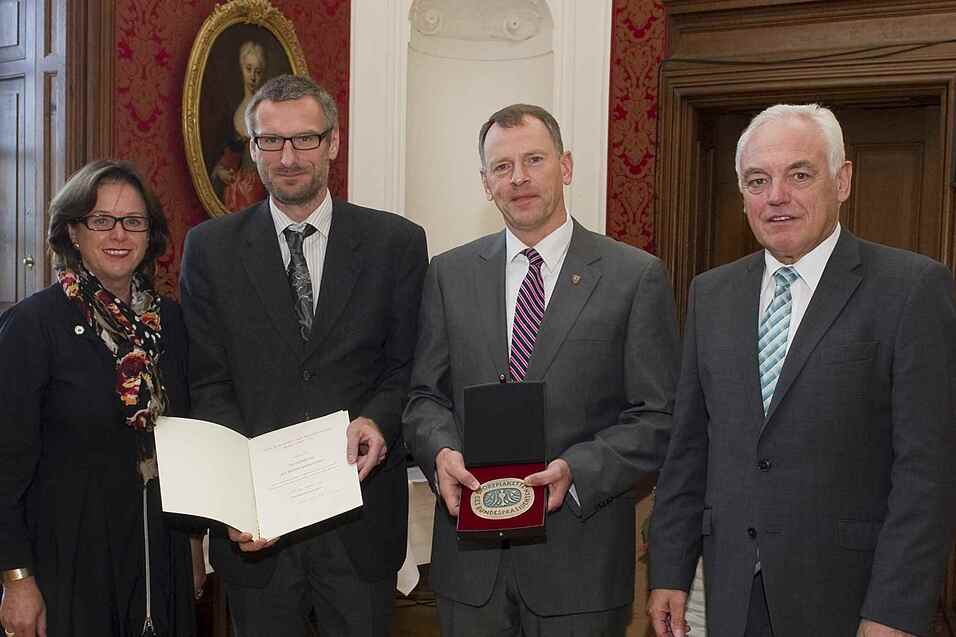 Vier Personen stehen in einem historischen Raum. Zwei halten Urkunde und Medaille, im Hintergrund ein Portr&auml;t.