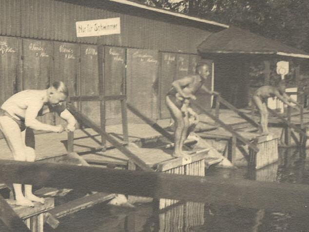 Vier Jungen in Badehosen springen von einem Holzsteg ins Wasser eines Sees, im Hintergrund eine Holzlaube.