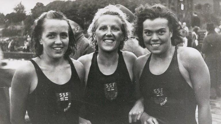 Drei Schwimmerinnen in Badekleidung l&auml;cheln zusammen im Freien, hinter ihnen sind Zuschauer am Ufer zu sehen.