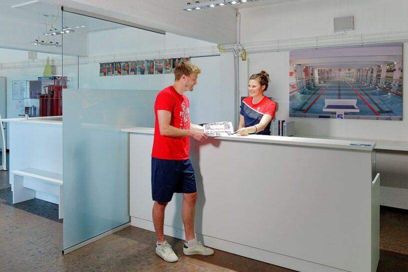 Mann im roten Shirt steht an einem Empfangstresen und spricht mit einer Frau in blauer Uniform. Moderne Sporteinrichtung im Hintergrund.