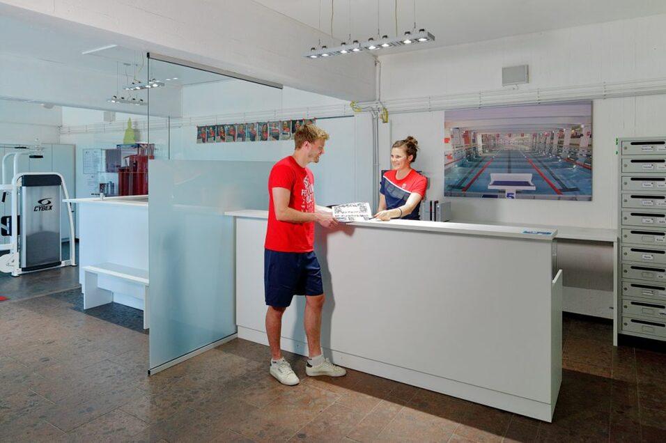 Mann im roten Shirt steht an einem Empfangstresen und spricht mit einer Frau in blauer Uniform. Moderne Sporteinrichtung im Hintergrund.