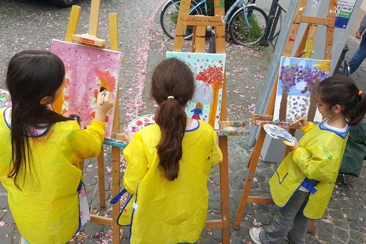 Drei Kinder in gelben Schutzsch&uuml;rzen malen an Staffeleien auf einer gepflasterten Stra&szlig;e, umgeben von Bl&uuml;tenbl&auml;ttern.