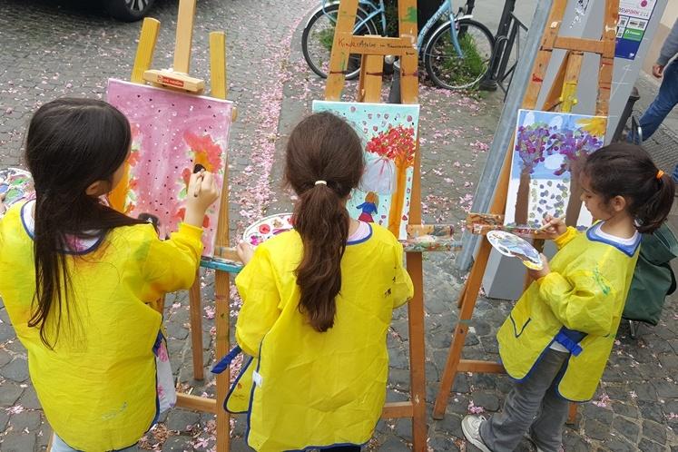 Drei Kinder in gelben Schutzsch&uuml;rzen malen an Staffeleien auf einer gepflasterten Stra&szlig;e, umgeben von Bl&uuml;tenbl&auml;ttern.