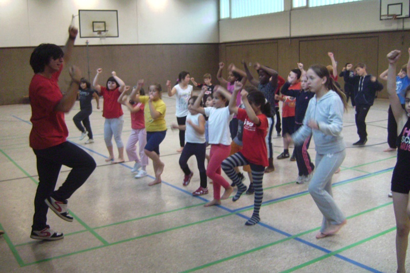 Gruppe von Kindern und Jugendlichen tanzt in einer Sporthalle zu den Anweisungen eines Lehrers in rotem Shirt.