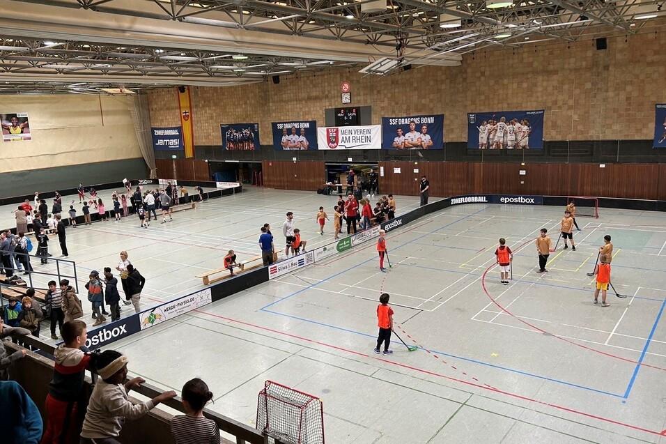 Sporthalle mit Gruppen von Kindern, die in rot-orangefarbenen Trikots Hockey spielen, Zuschauer am Rand.