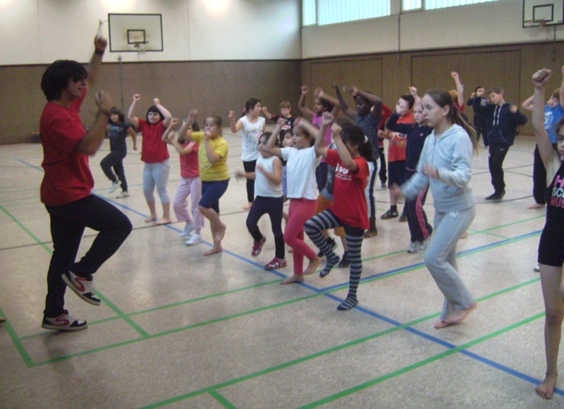 Gruppe von Kindern und Jugendlichen tanzt in einer Sporthalle zu den Anweisungen eines Lehrers in rotem Shirt.