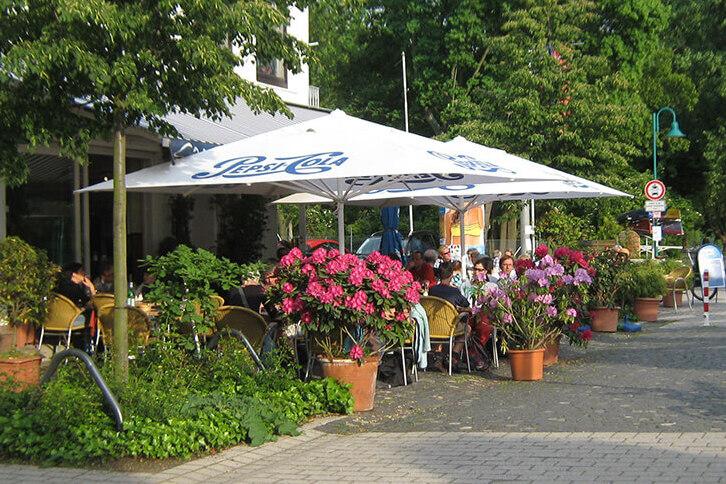 Szenenansicht eines Caf&eacute;s mit Sonnenschirmen, Blumen und G&auml;sten an einem gepflasterten Weg. Fahrr&auml;der sind im Hintergrund.