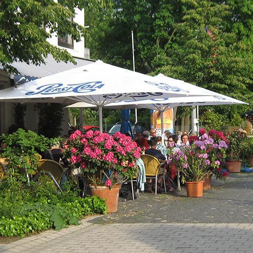 Szenenansicht eines Caf&eacute;s mit Sonnenschirmen, Blumen und G&auml;sten an einem gepflasterten Weg. Fahrr&auml;der sind im Hintergrund.