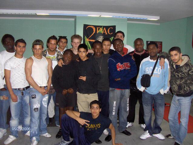 Gruppe junger M&auml;nner posiert gemeinsam in einem Raum mit &bdquo;2Pac&ldquo;-Plakat im Hintergrund.