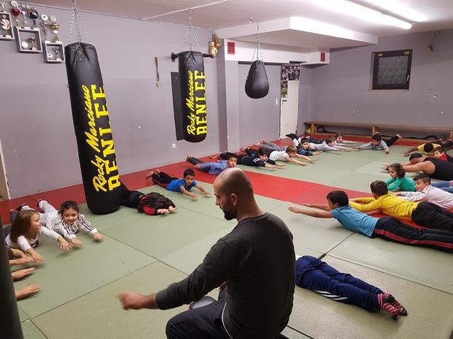 Gruppe von Kindern und Jugendlichen beim Training in einer Sporthalle, liegend auf Matten mit einem Trainer in der Mitte.