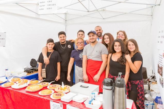Gruppe von acht Personen l&auml;chelt vor einem Festzelt, umgeben von verschiedenen Kuchen auf einem roten Tisch.