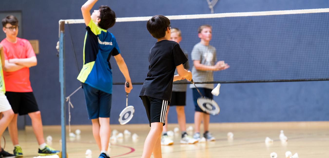 Gruppe von Kindern beim Badmintontraining in einer Sporthalle, mit Schlägern und Federbällen auf dem Boden.