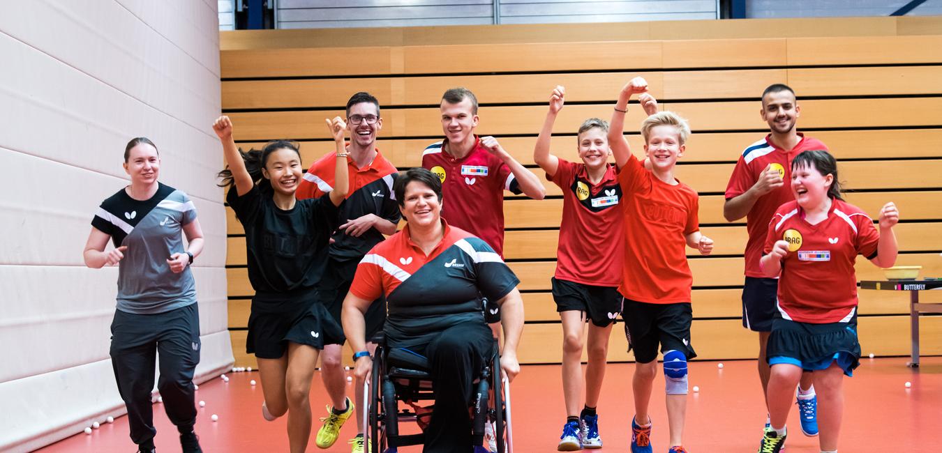 Gruppe von Tischtennisspielern in Sportkleidung läuft lächelnd und jubelnd auf rotem Boden in einer Sporthalle.
