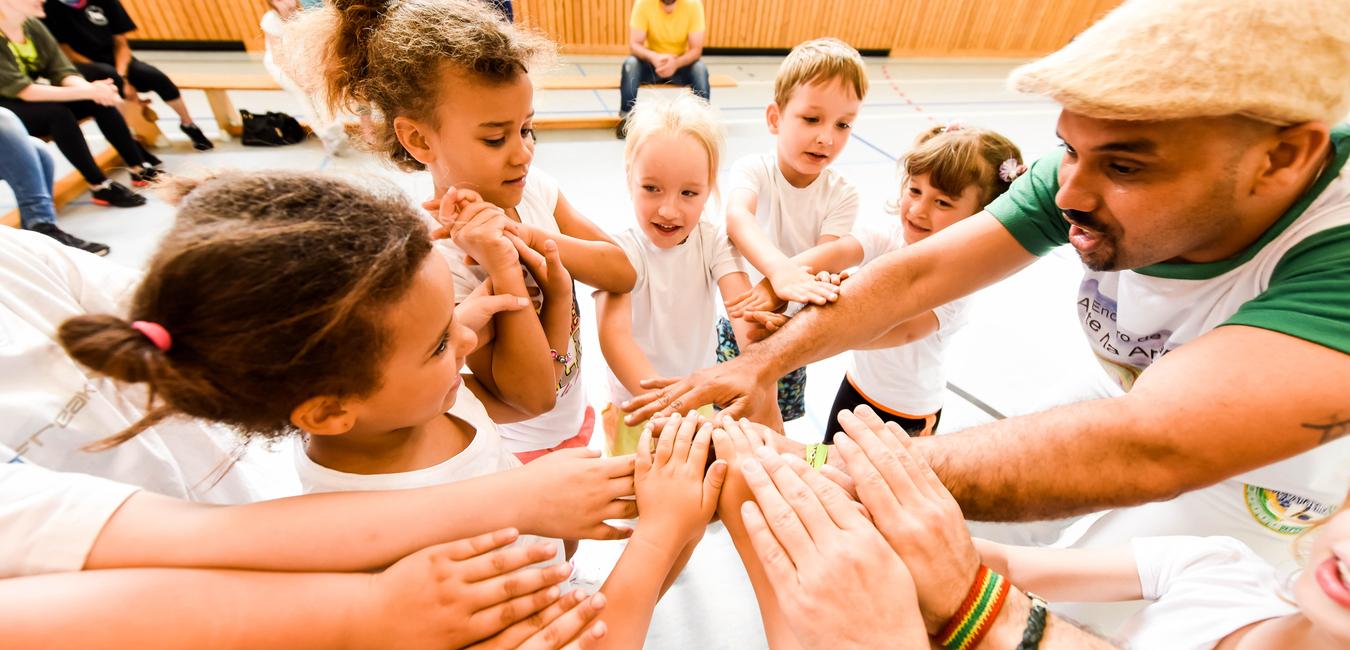 Gruppenspiel mit Kindern, die ihre Hände zum gemeinsamen Aufeinandertreffen heben, unter Anleitung eines Erwachsenen.
