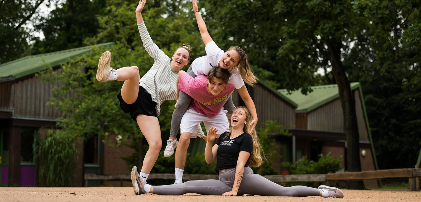 Vier junge Frauen posieren fröhlich auf einer sandigen Fläche, eine sitzt im Spagat, während die anderen sie umarmen.