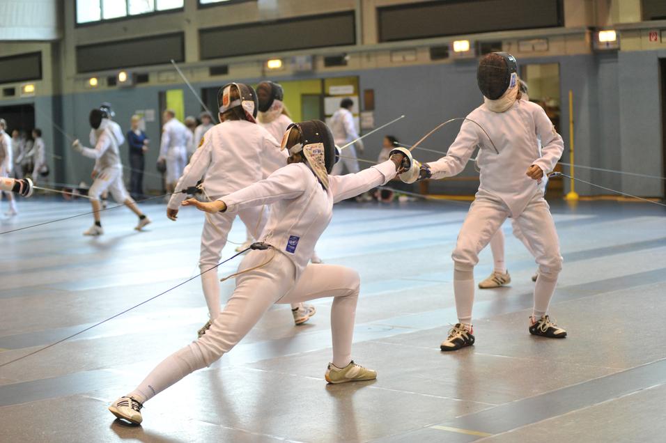 Zwei Fechter in Kampfpose in einem Sportzentrum, umgeben von weiteren Fechtern und Zuschauern.