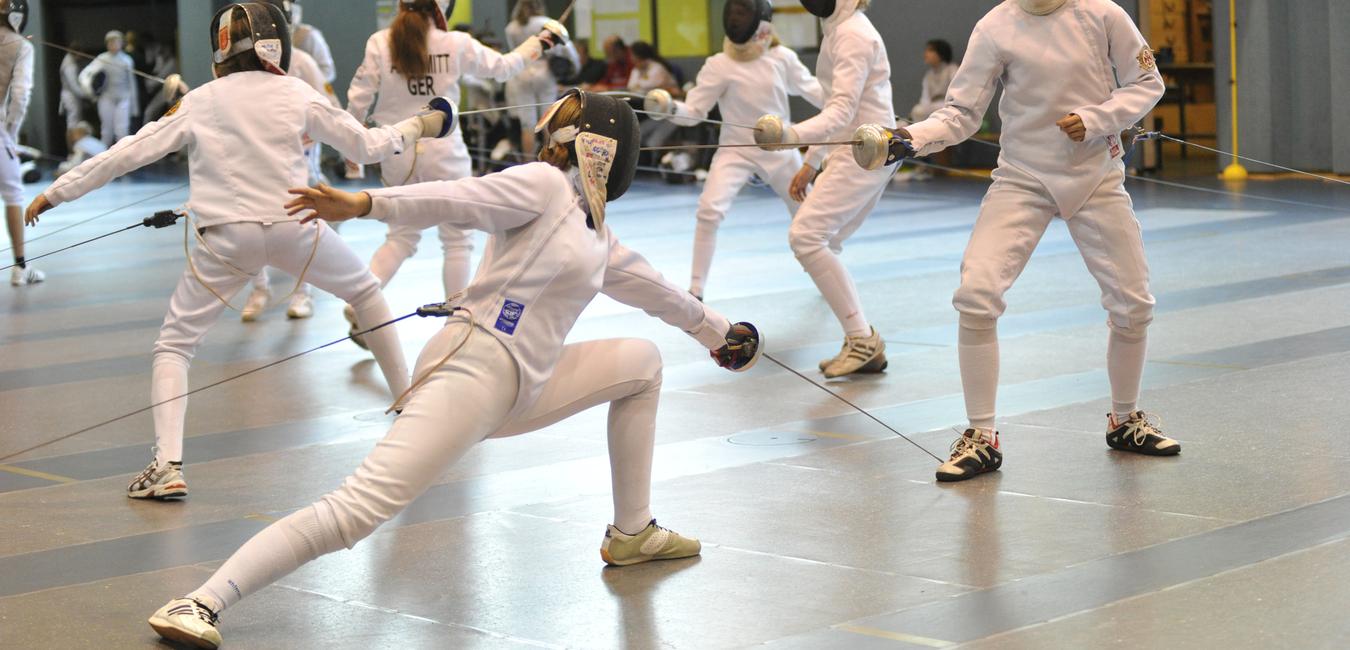 Fünf Fechterinnen in weißer Schutzkleidung trainieren in einer Sporthalle, während sie ihre Säbel einsetzen.