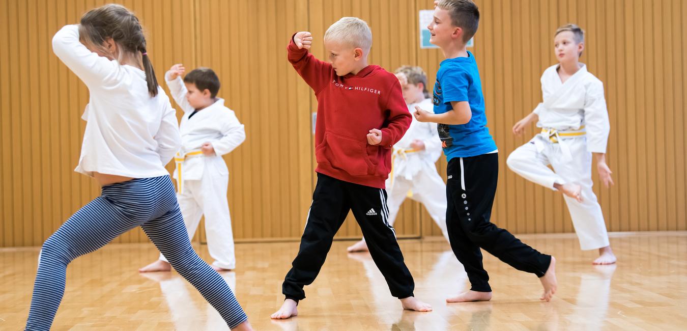 Sechs Kinder üben in einem Sportraum Kampfsportbewegungen in weißer und bunter Kleidung auf einem Holzfußboden.