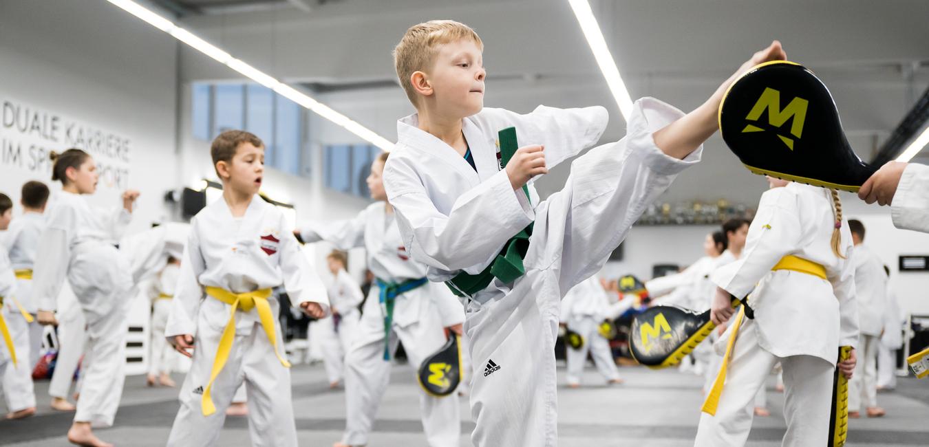 Eine Gruppe von Kindern in Karate-Uniformen übt in einem Trainingsraum, ein Junge führt einen Kick aus.