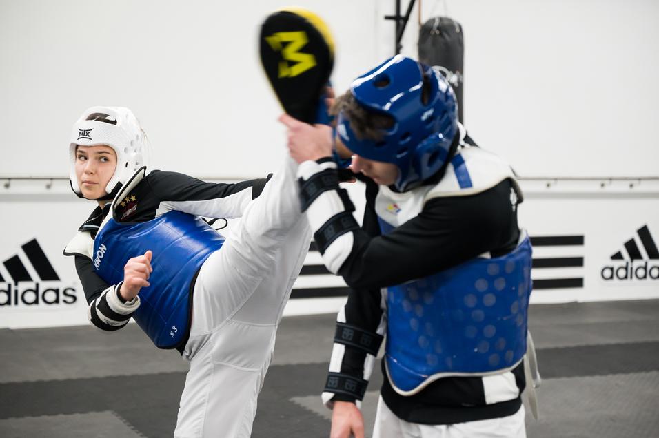 Zwei Kampfkämpfer im Taekwondo-Training; eine Person führt einen hohen Kick aus, beide tragen Schutzausrüstung.