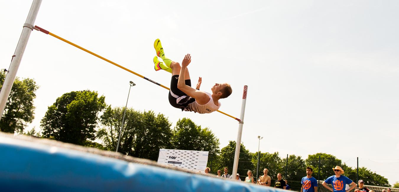 Springer beim Hochsprung über eine gelbe Lattenstange, im Hintergrund Zuschauer und grünes Umfeld.