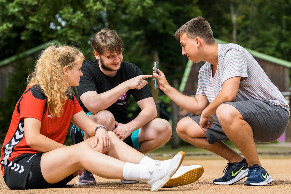 Drei Jugendliche sitzen auf einem Sandplatz und betrachten gemeinsam ein Smartphone. Bäume und Hütten im Hintergrund.