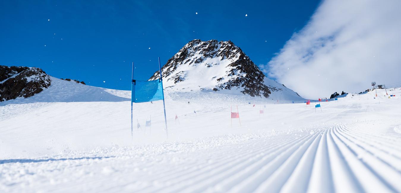 Schneebedeckte Piste mit blauen und roten Toren, im Hintergrund ein schneebedeckter Berg unter blauem Himmel.