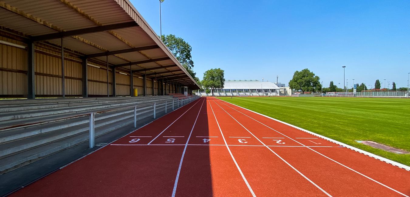 Leere Laufbahn mit roten Bahnen und Tribüne unter einem klaren blauen Himmel, neben einem Kunstrasenfeld.
