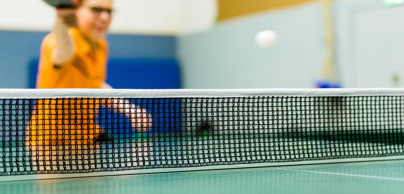 Tischtennisschläger in der Hand eines Spielers, im Hintergrund ein Ball über das Netz fliegend auf einer grünen Tischtennisplatte.