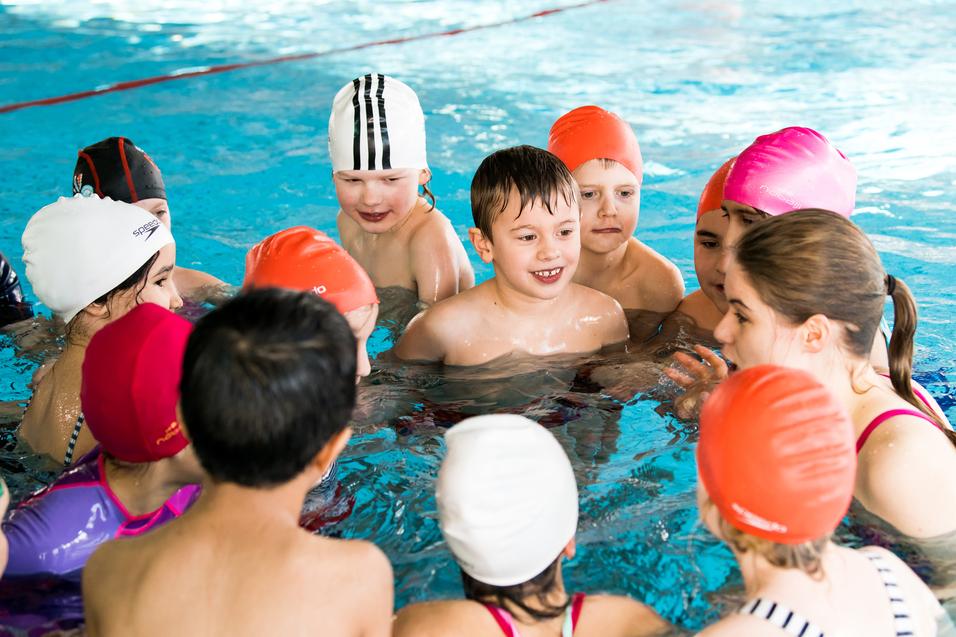 Gruppe von Kindern im Schwimmbecken, die sich um eine Trainerin versammeln und miteinander kommunizieren.