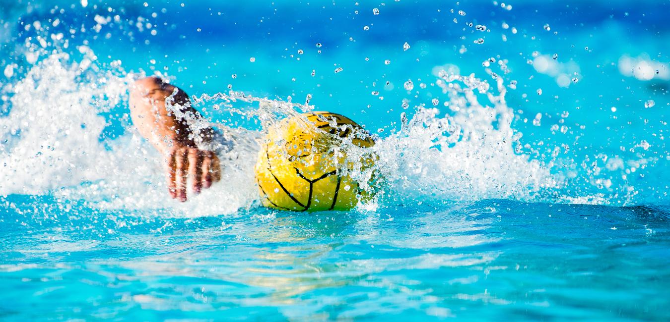 Hand greift nach einem gelben Wasserball, der im spritzenden Wasser eines Schwimmbeckens schwimmt.